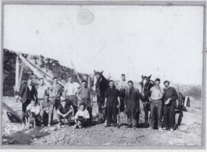 Grayscale picture of quarry workers standing and sitting in a quarry for a photo shoot - with their horses and a dog.